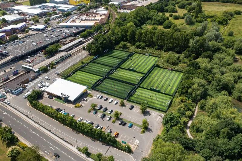Aerial view of a sports complex with three football pitches with striped grass markings, parking area, and pavilion building surrounded by trees and roads.