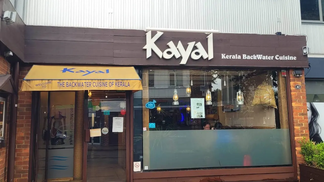 Storefront of Kayal Kerala BackWater Cuisine restaurant with brown and yellow signage, large windows, and hanging lanterns visible inside.