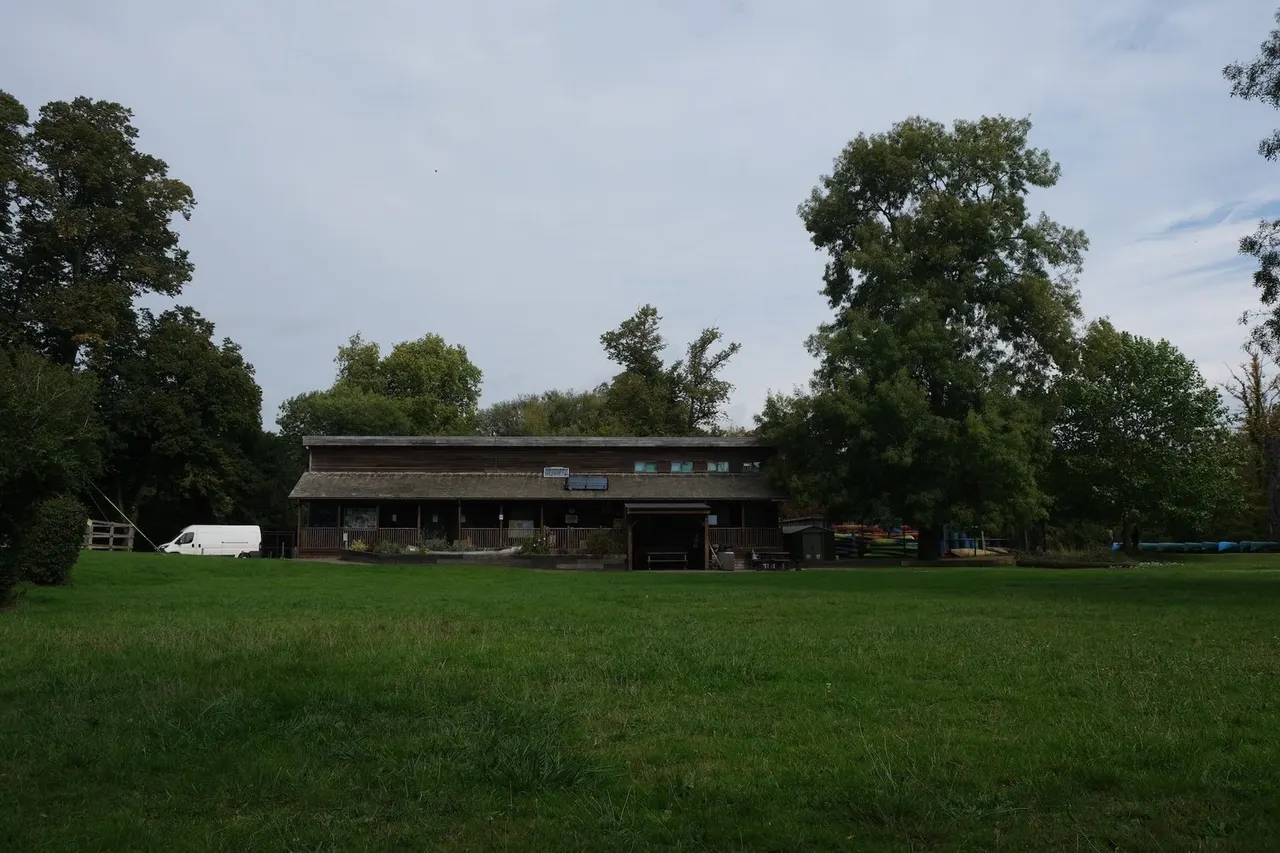 A brown wooden building with a covered porch sits across a large green field, surrounded by trees.