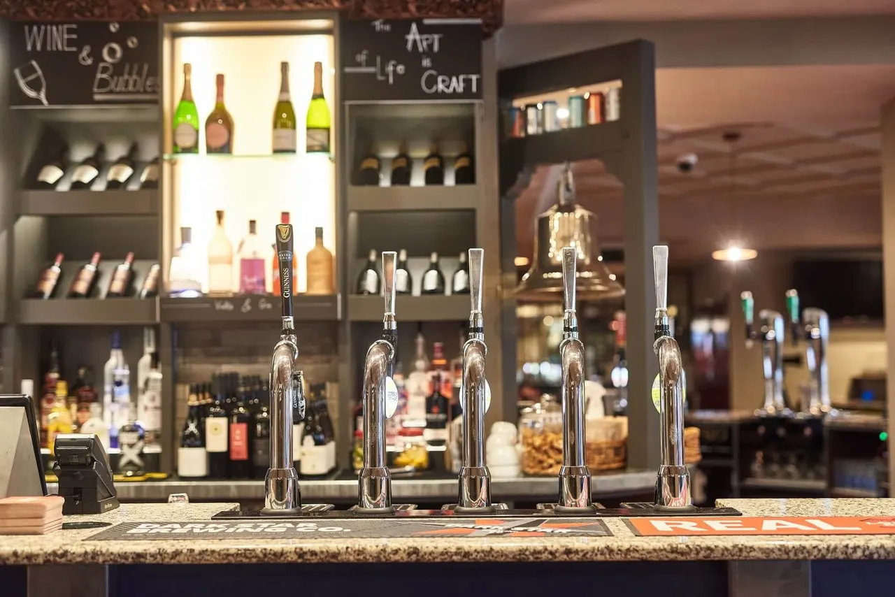 Bar counter with five gleaming beer taps, shelving displaying wine and spirit bottles behind, and signage for wine and craft offerings.
