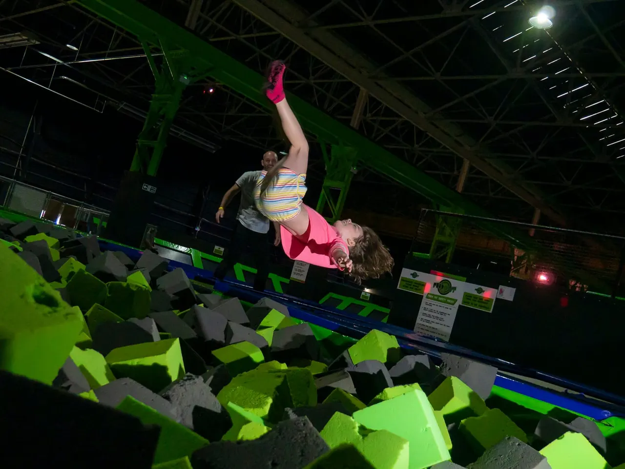 Child performing a backflip on a trampoline at an indoor arena with neon green foam pits.