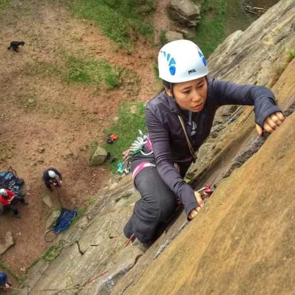 A person in a climbing helmet ascends a steep rock face with safety equipment, viewed from above with spotters on the ground below.
