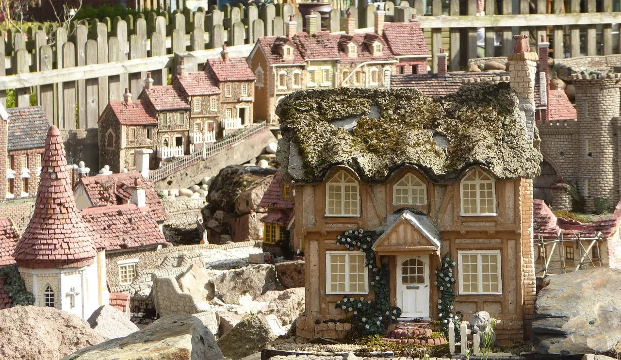 Miniature model village with detailed stone cottages, red-tiled roofs, and moss-covered buildings arranged on a landscape with rocks and fencing.