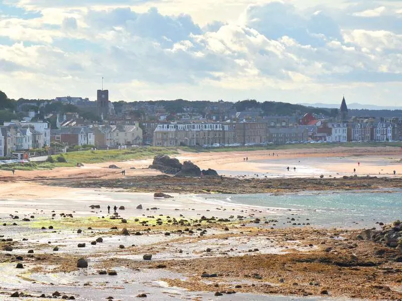 Rocky foreshore and sandy beach with scattered visitors, backed by a seaside town with historic buildings under cloudy sky.