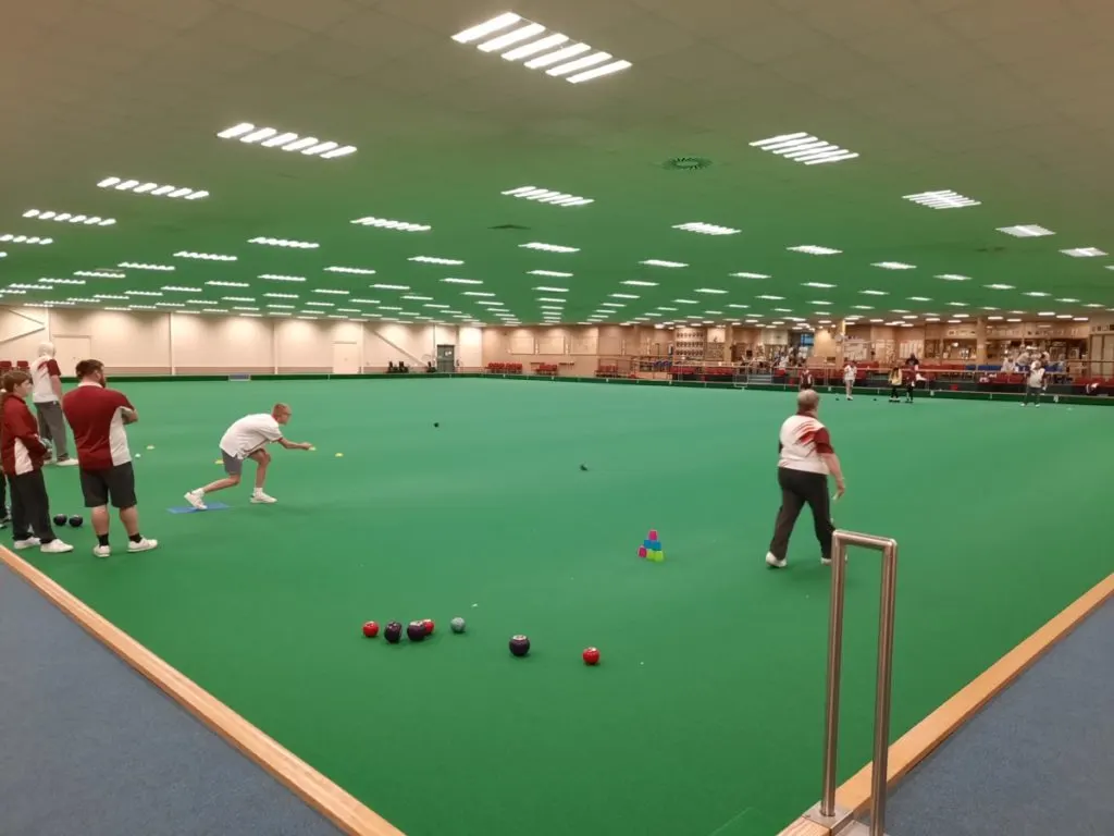 Players competing in lawn bowls on an indoor green, with spectators watching from the sides.