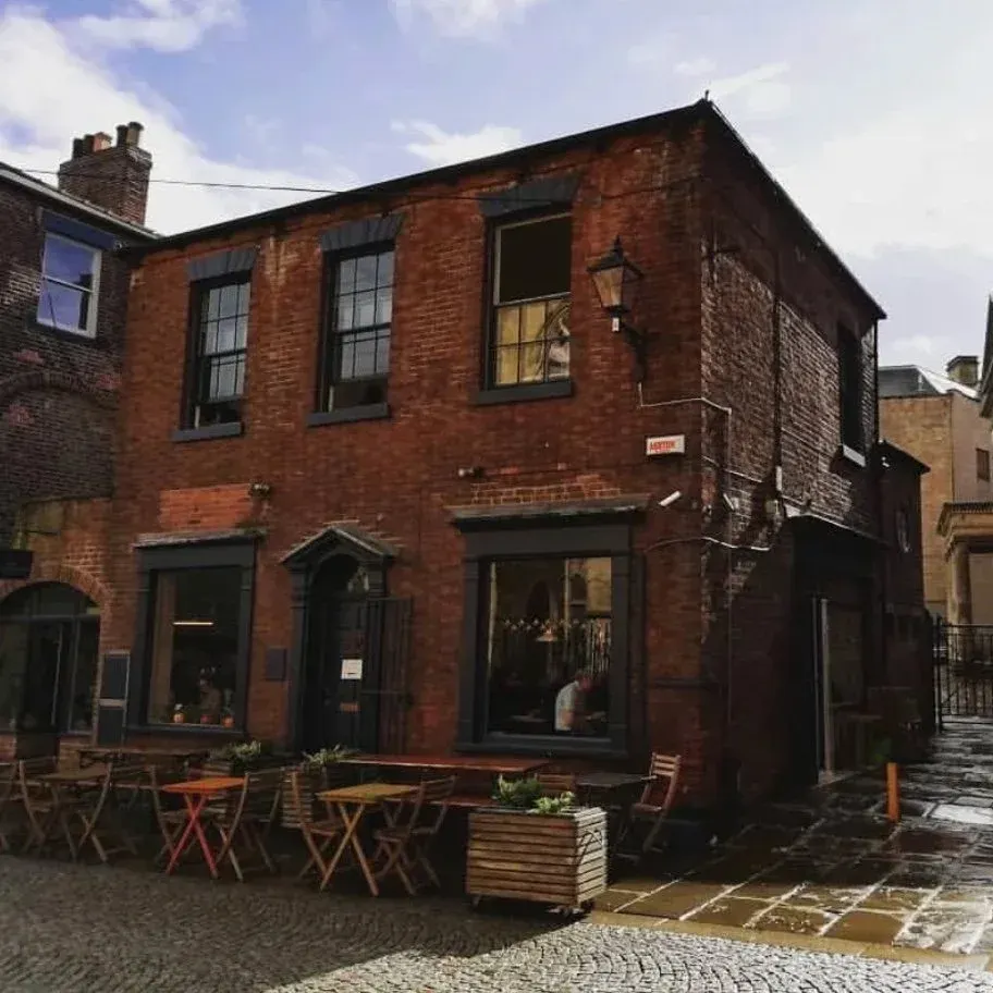 Red brick building with outdoor seating area featuring wooden tables and chairs on a cobbled courtyard.