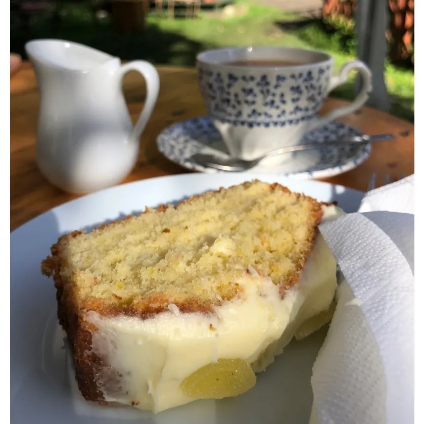 Slice of Victoria sponge cake with cream and jam filling on a plate, with teacup and milk jug on outdoor table.