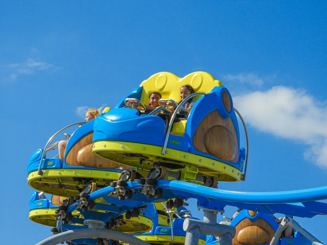 Families riding a blue and yellow roller coaster with arms raised against a clear blue sky.