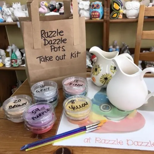 A pottery painting take-out kit displayed on a table with labelled paint containers, brushes, white ceramic pieces, and shelves of supplies in the background.