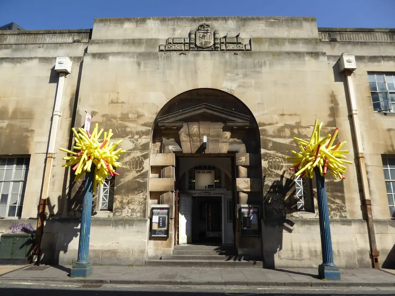 Historic stone police station building with large arched entrance, decorated with bright yellow sculptural installations flanking the doorway.
