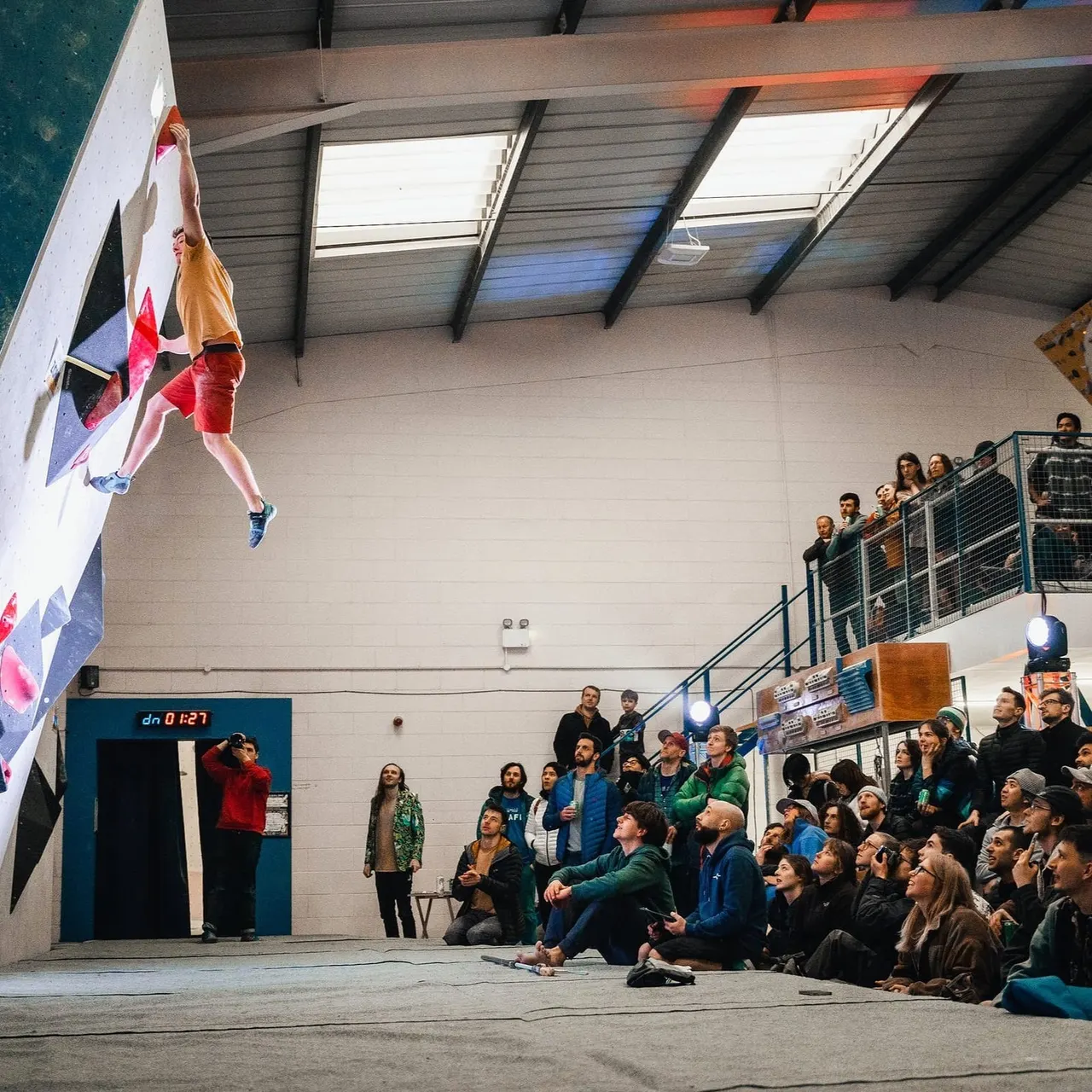 Person indoor rock climbing on a high wall whilst spectators watch from below in a sports hall.