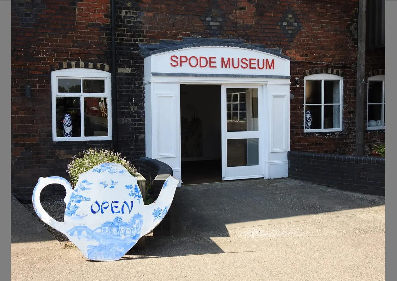 White storefront building with "Spode Museum" signage and a large decorative teapot sign marked "OPEN" outside against red brick wall.