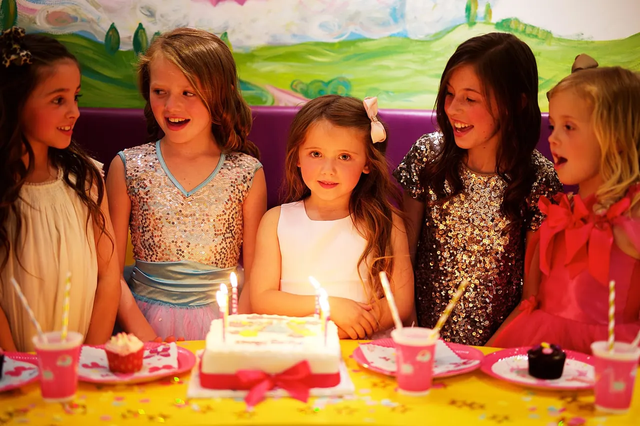 A group of five children gathered around a birthday cake with lit candles at a colourful party table.