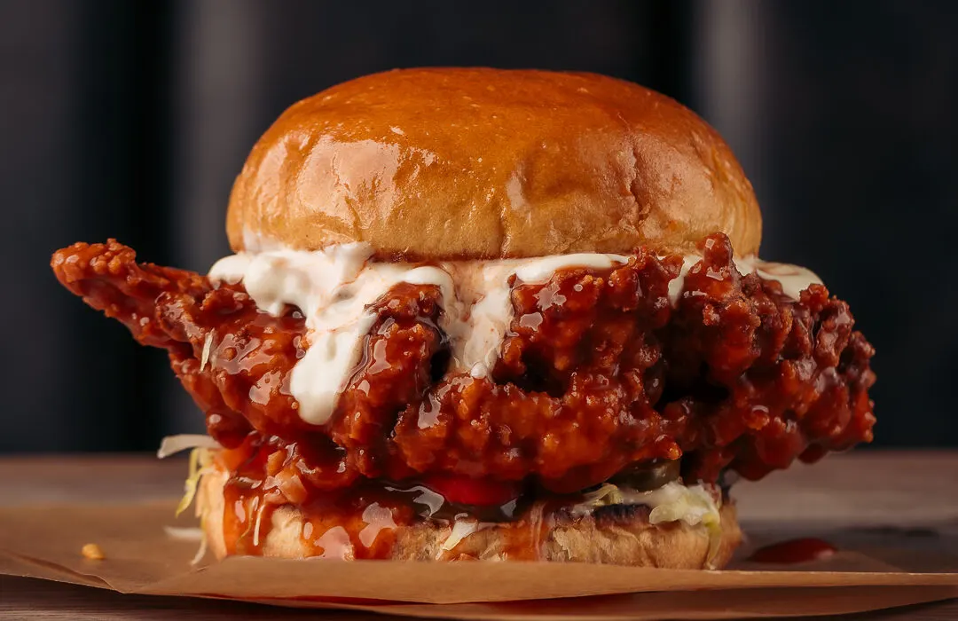Crispy fried chicken sandwich with spicy glaze and white sauce on a toasted bun.