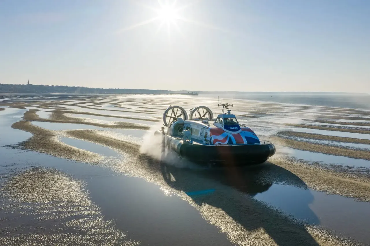 A blue and red hovercraft with large propeller fans travels across shallow water and mudflats on a sunny day.