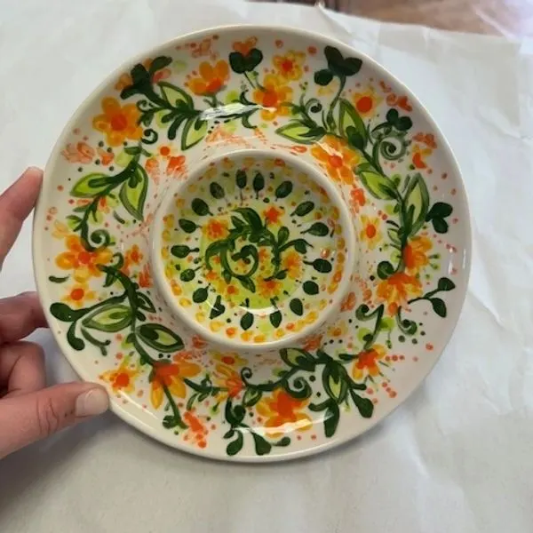 Hand holding a decorative ceramic dish with floral and leaf patterns in orange, green and yellow.