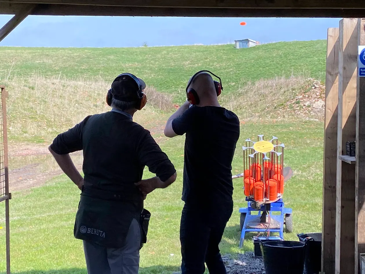 Two people wearing headphones and dark clothing stand at a shooting range, aiming at clay targets on a blue machine in an open field.