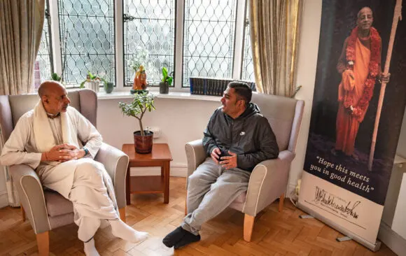 Two men sit in cream armchairs conversing in a bright, plant-filled room with latticed windows and religious artwork on the walls.