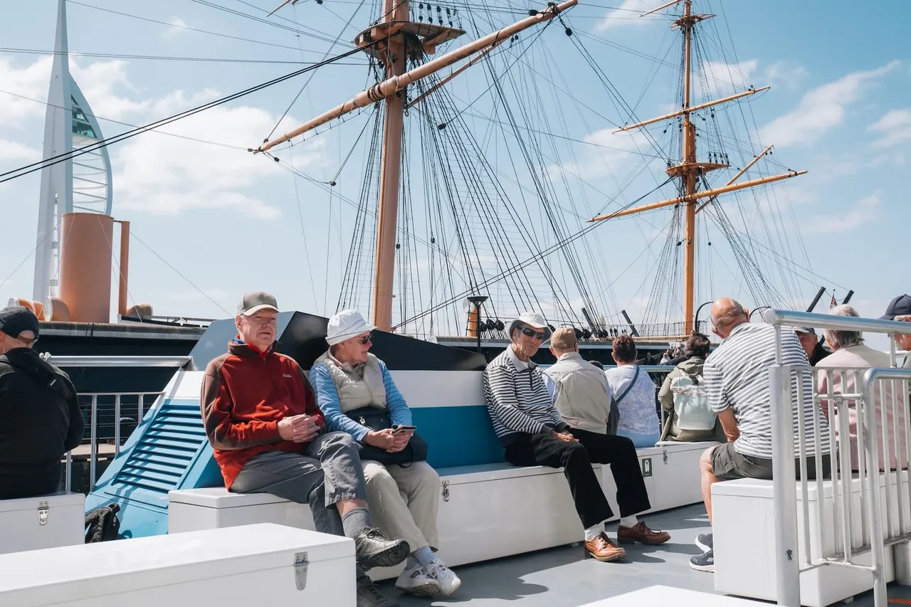 Passengers seated and standing on the deck of a historic tall ship with rigging visible against a blue sky.