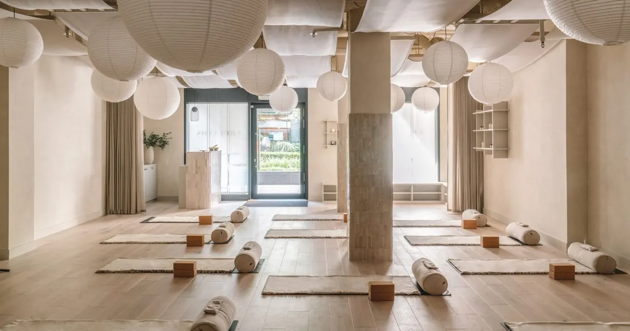 Spacious, minimalist yoga studio with yoga mats arranged in rows, wooden bolsters, paper lantern lighting, and large windows overlooking gardens.