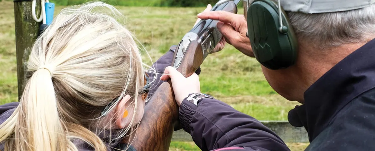 Two people clay pigeon shooting, one aiming a shotgun whilst wearing headphones.