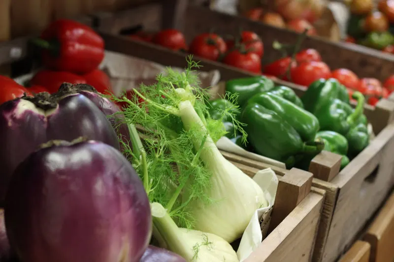 Fresh produce displayed in wooden crates including aubergines, fennel, green peppers and tomatoes.