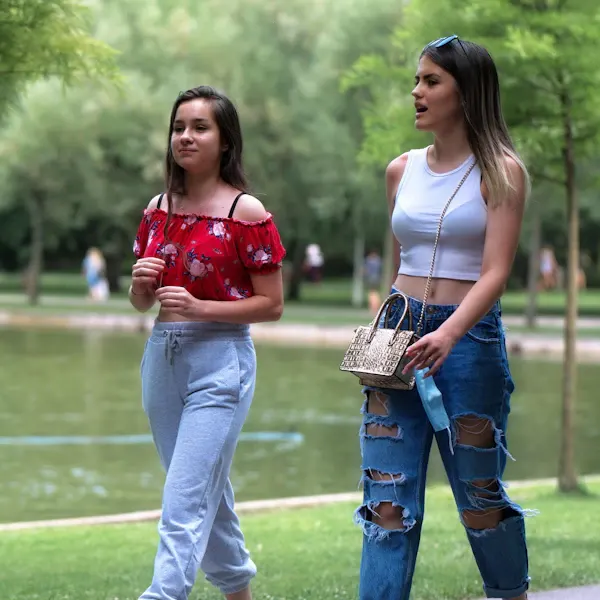 Two young women walking beside a lake in a park with trees and other visitors visible in the background.