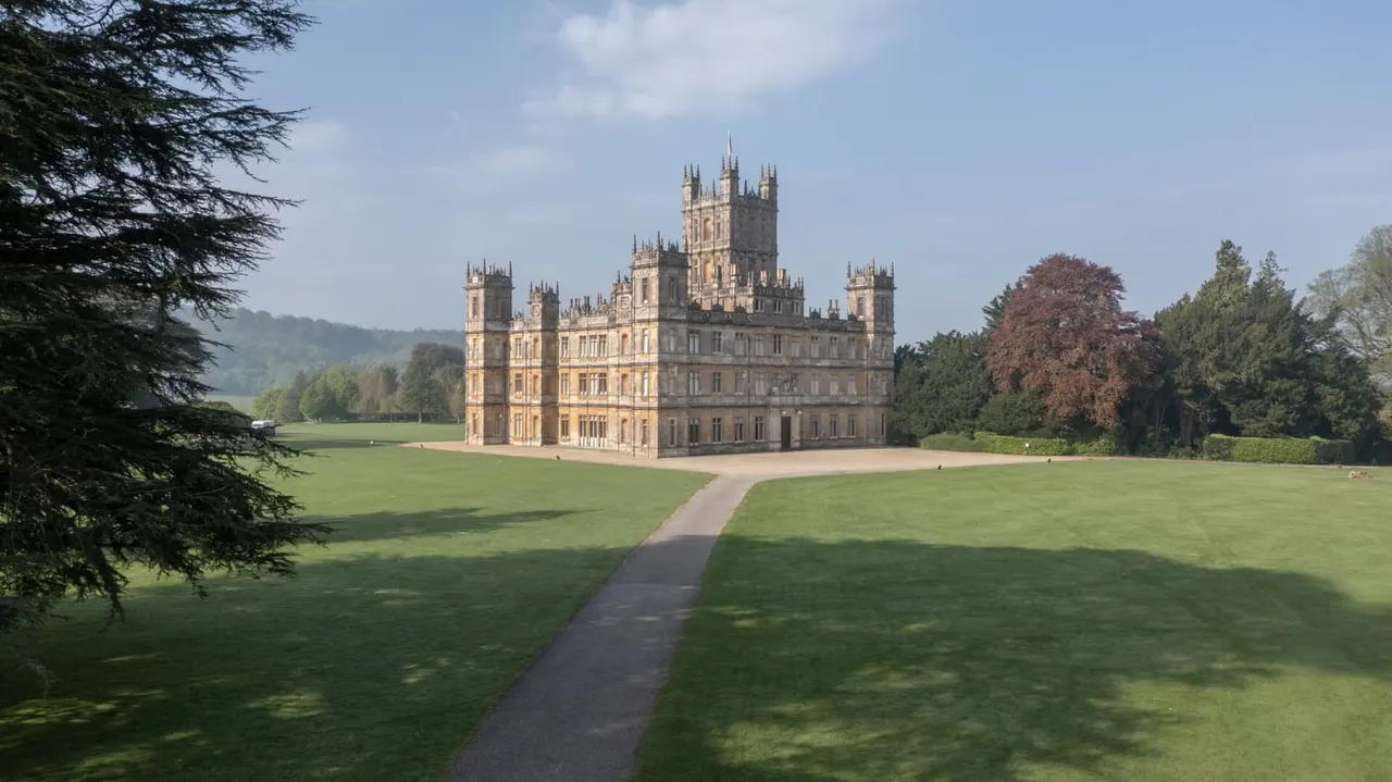 A grand stone castle with multiple turrets and spires sits in an expansive parkland with manicured lawns, mature trees and a pathway leading to its entrance.