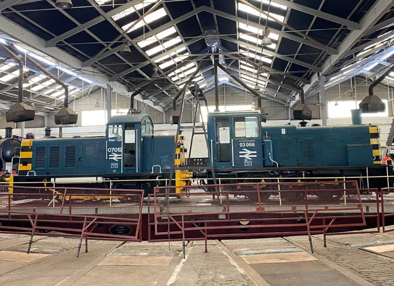 Two preserved blue diesel locomotives on display inside a railway shed with a glazed roof and safety fencing.