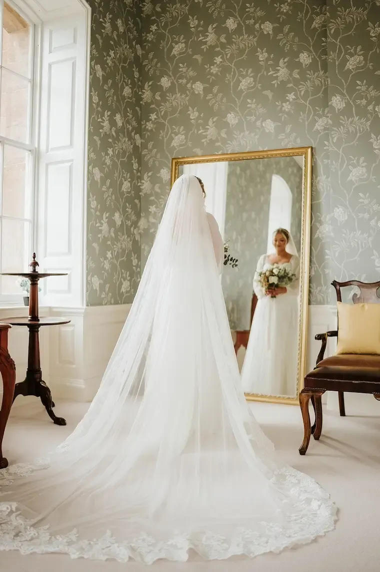 Bride in white wedding gown and veil viewing herself in a large mirror in an elegant period room with floral wallpaper.