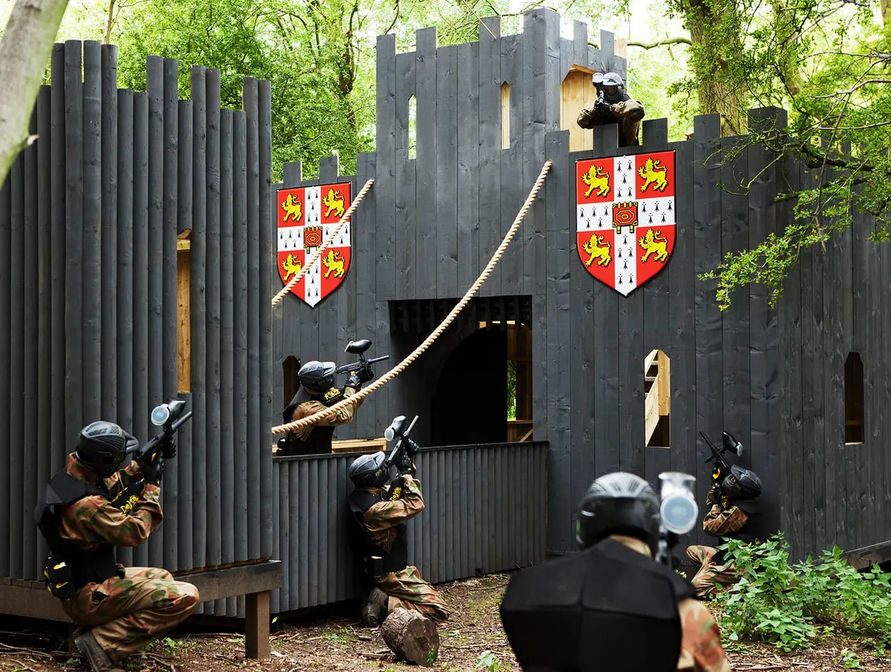 Players in combat gear and masks aim paintball guns at a medieval-style castle structure during an outdoor paintball game.