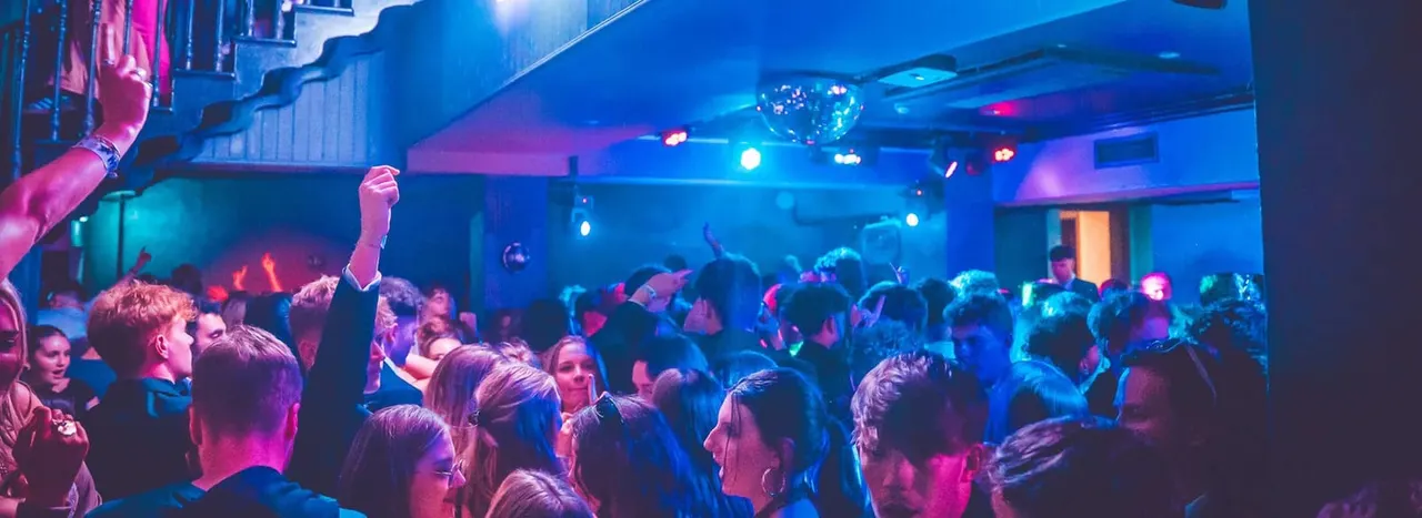 Crowded dance floor at night club with neon blue and pink lighting, dancers enjoying music and raising their arms.