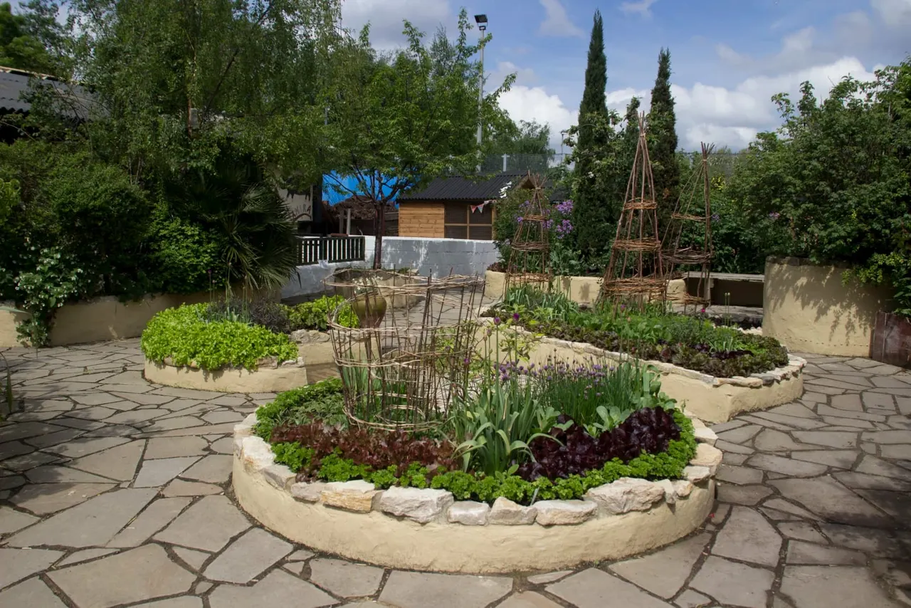 Raised beds with vegetables and herbs in a paved garden area surrounded by trees and farm buildings.