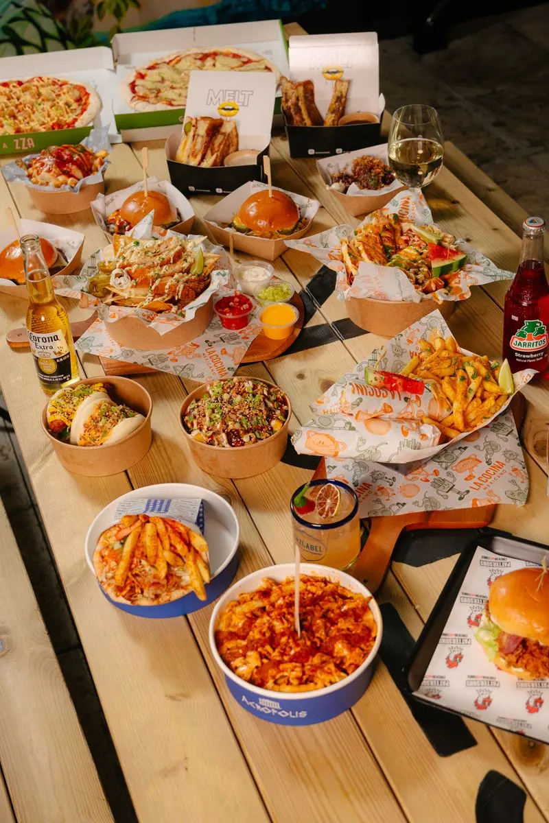Overhead view of a wooden table spread with pizzas, burgers, hot dogs, fries, salads and drinks at a casual dining venue.
