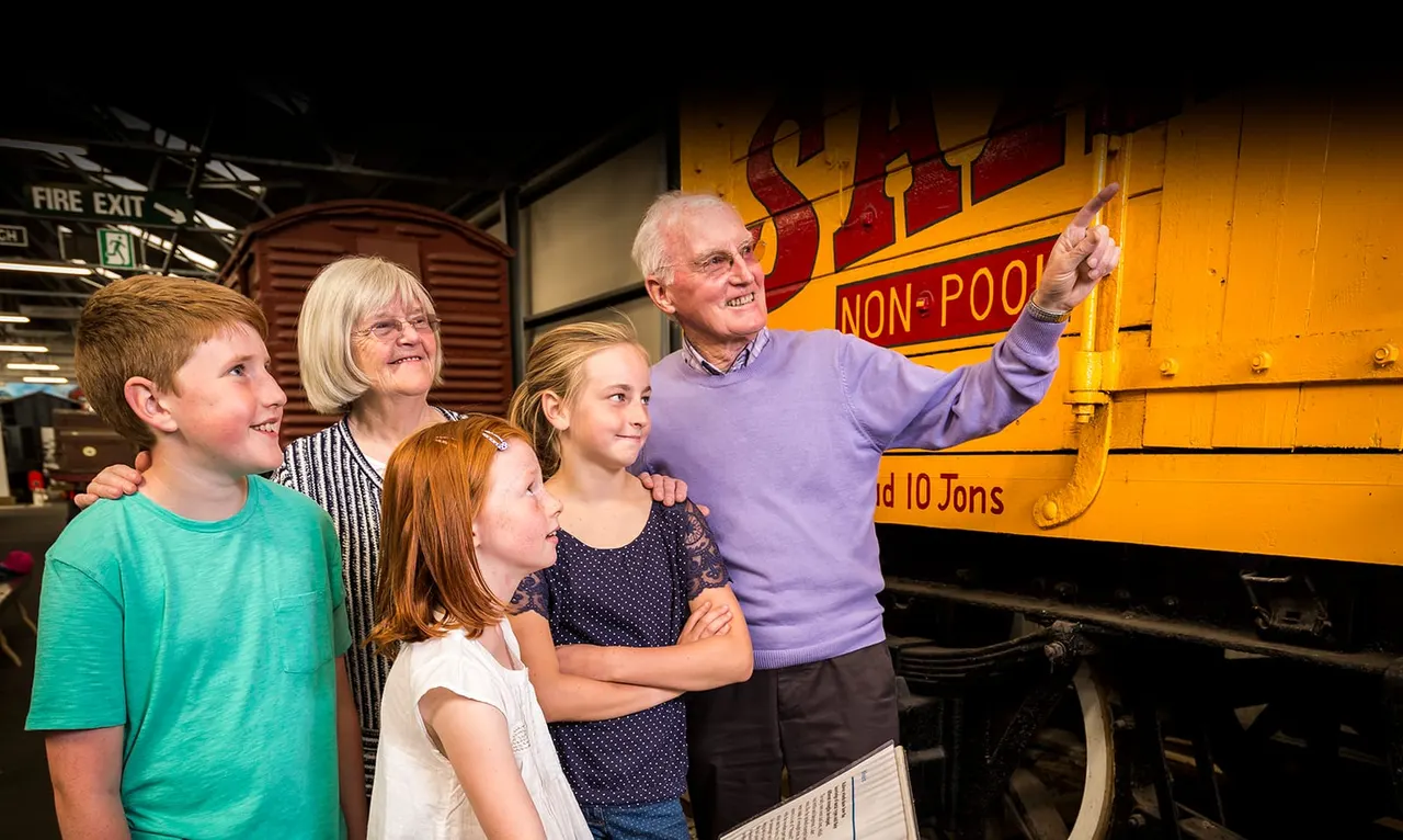 An elderly man and woman with three children stand beside a large yellow heritage railway locomotive, smiling and looking at the engine.