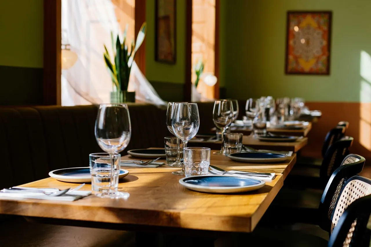 Formal dining table set with white plates with blue trim, wine glasses, water glasses, cutlery and napkins in a bright room with green walls and flower arrangements.