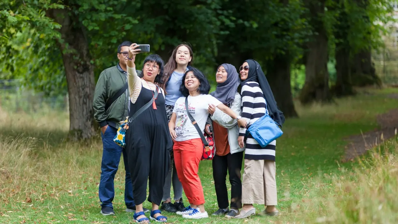 A group of visitors takes a selfie together on a grassy woodland path lined with trees.