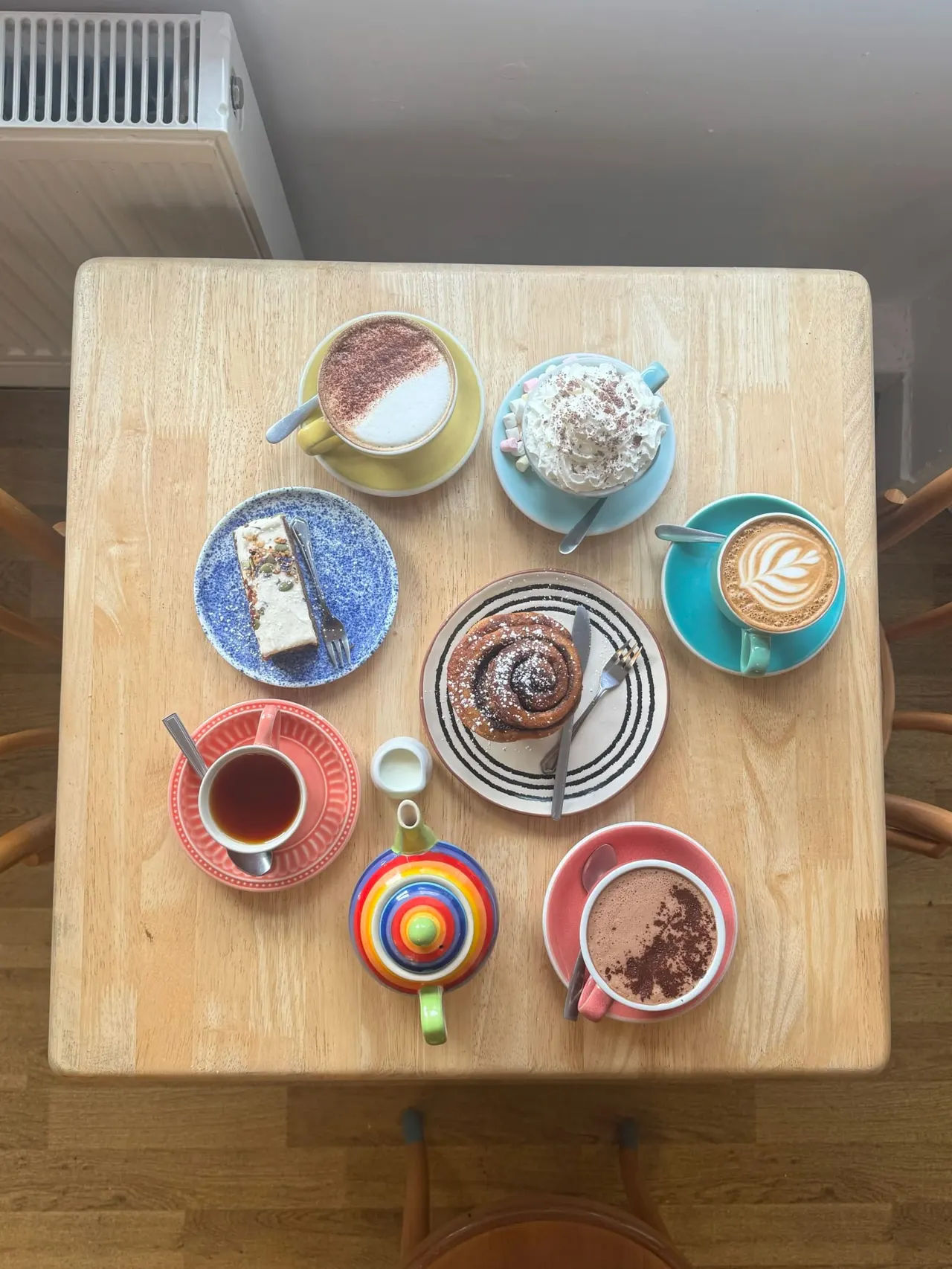 Overhead view of a wooden table displaying an assortment of colourful cups and plates with hot beverages, cakes and desserts.