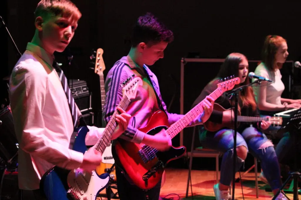 Young musicians performing on stage with electric guitars and microphone under coloured stage lighting.