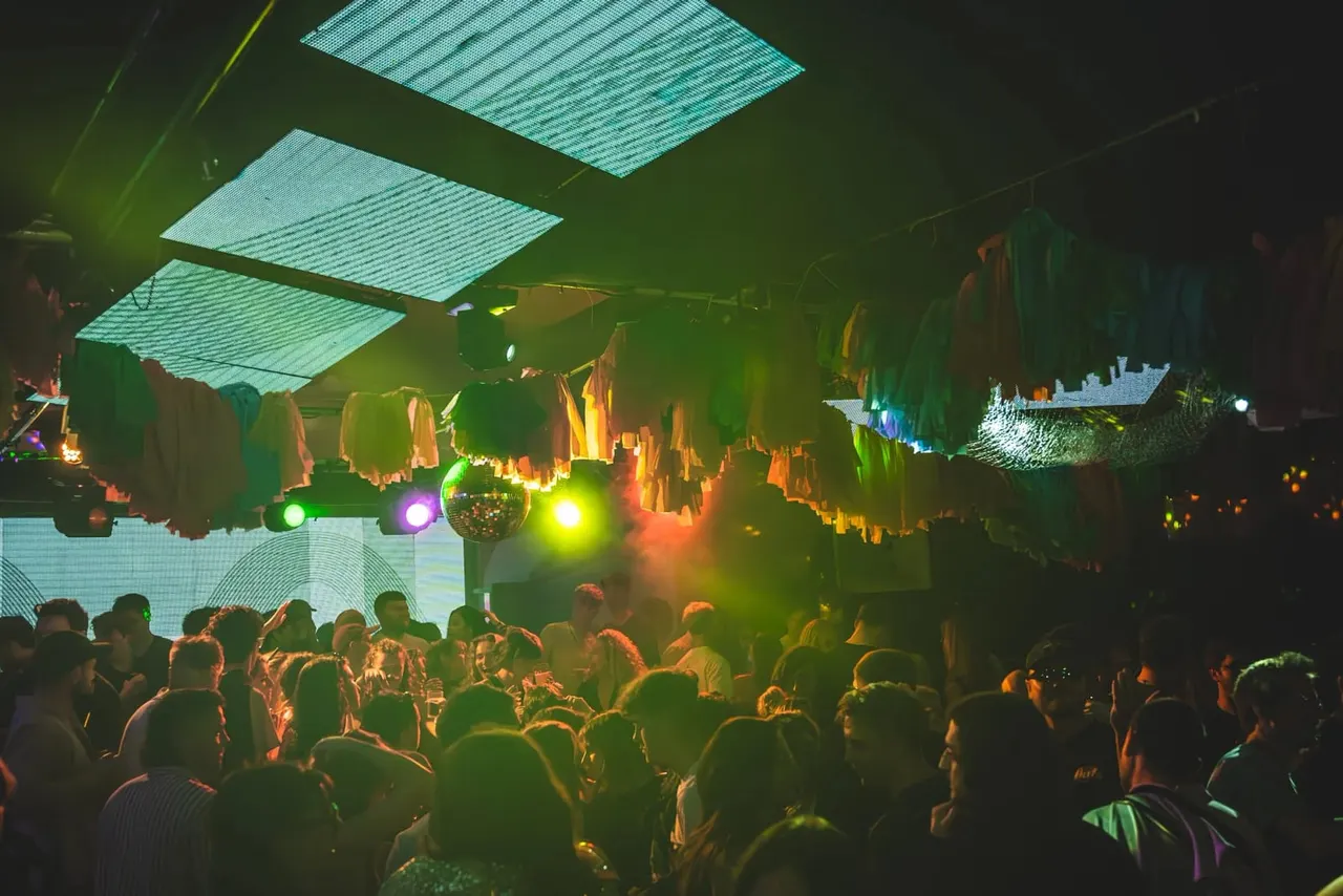 Crowded nightclub dance floor lit by coloured lights and laser displays, with suspended clothing and decorative elements overhead.