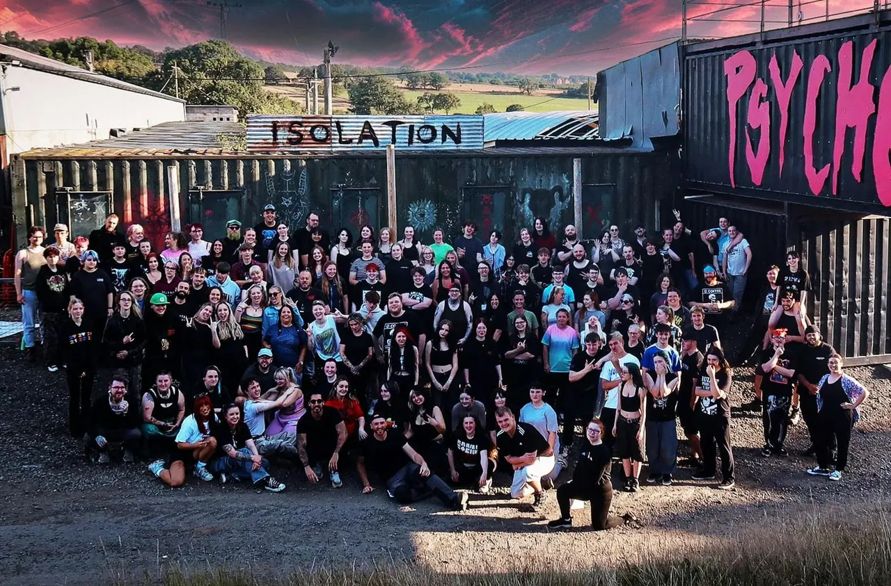 Large group of visitors posing together outside industrial-style buildings with graffiti art and pink sunset sky.