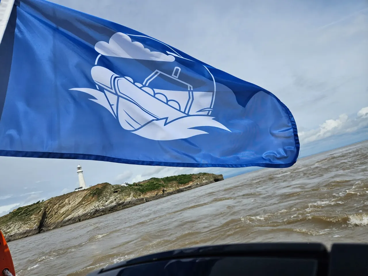 Blue flag with boat illustration flying from a vessel, with rocky headland and lighthouse visible across the water.