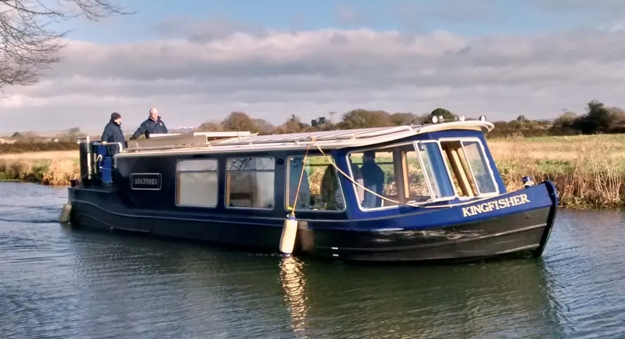 Blue and black narrowboat named Kingfisher moored on a canal with two people on the roof, rural landscape beyond.