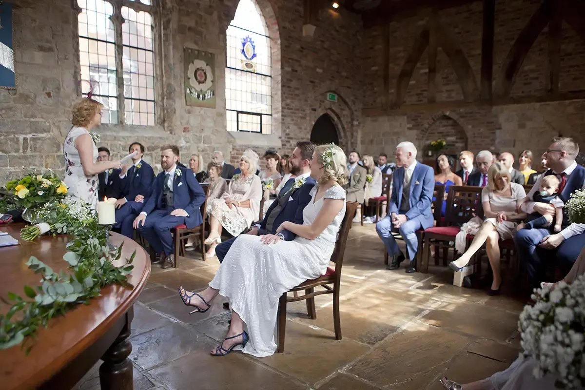 Wedding ceremony in a stone-walled historic hall with guests seated facing a bride and groom at the altar.