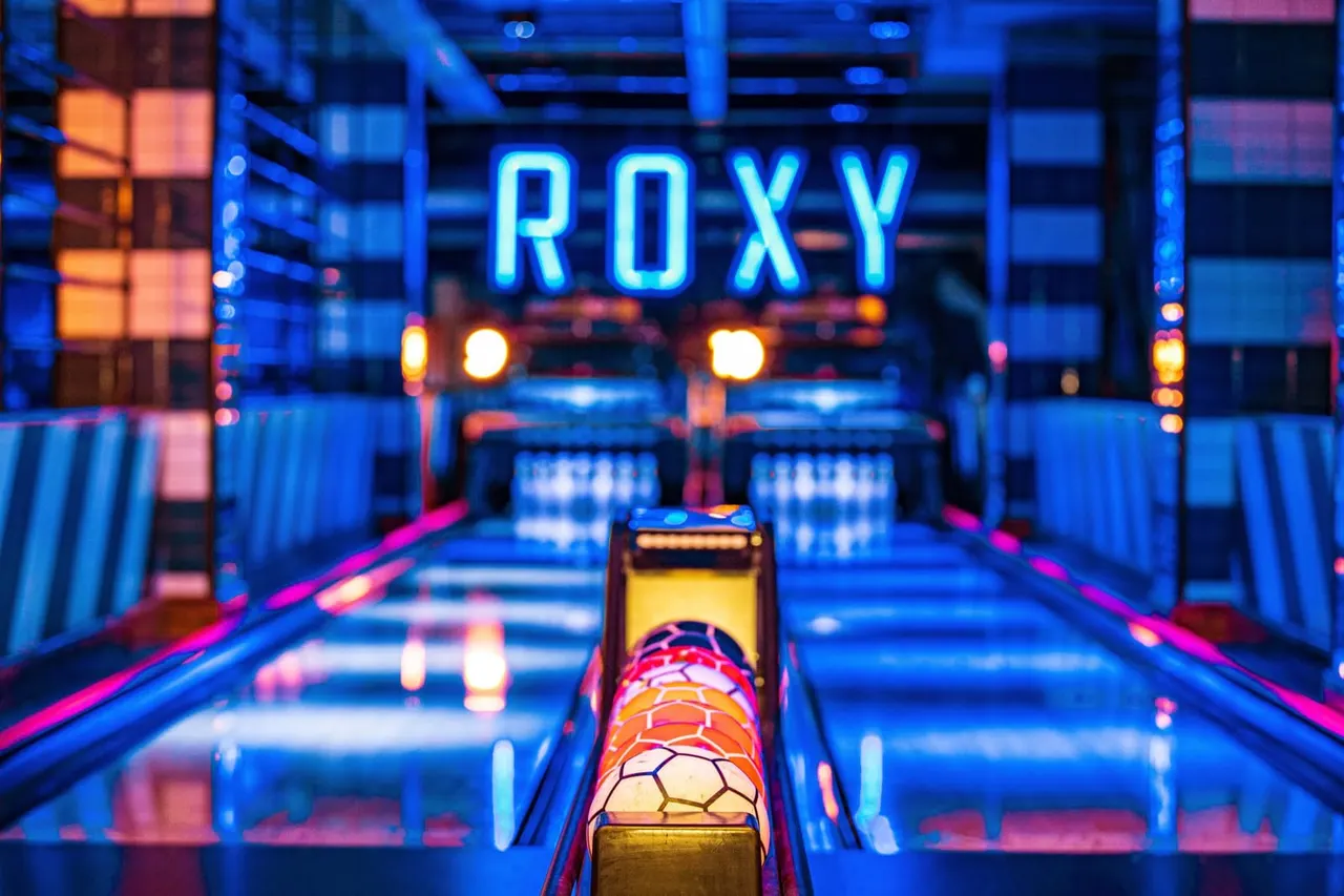 Brightly lit bowling lane with a coloured ball in the foreground and the Roxy neon sign glowing above.