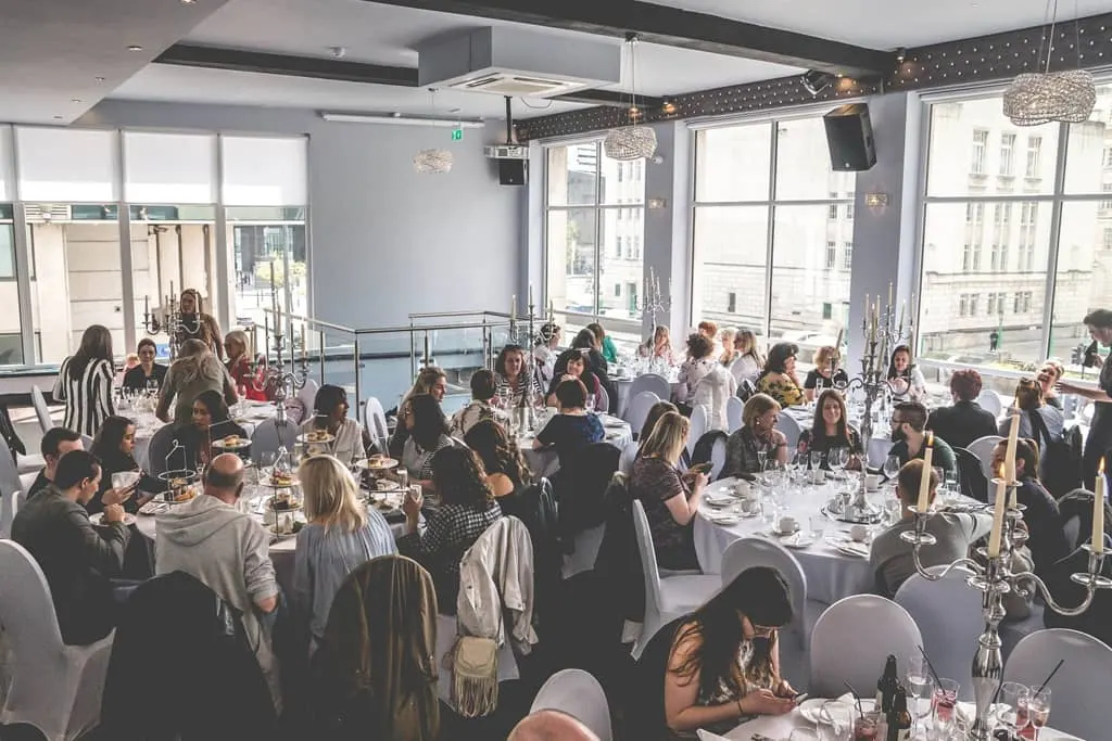 A large group of people dine together at tables in a contemporary restaurant with floor-to-ceiling windows overlooking a city.