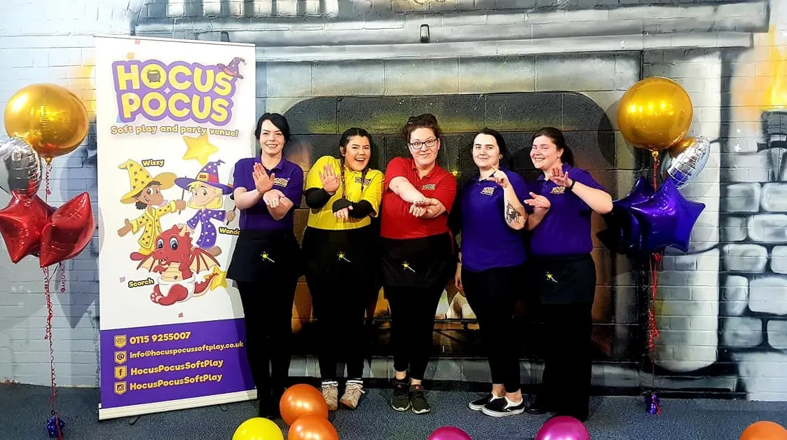 Five staff members in colourful tops pose together in front of a Hocus Pocus branded sign at a soft play venue.