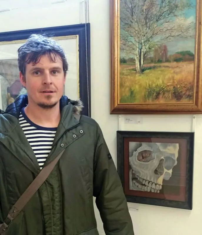 Man in olive jacket and striped shirt standing indoors next to framed artworks on a wall.