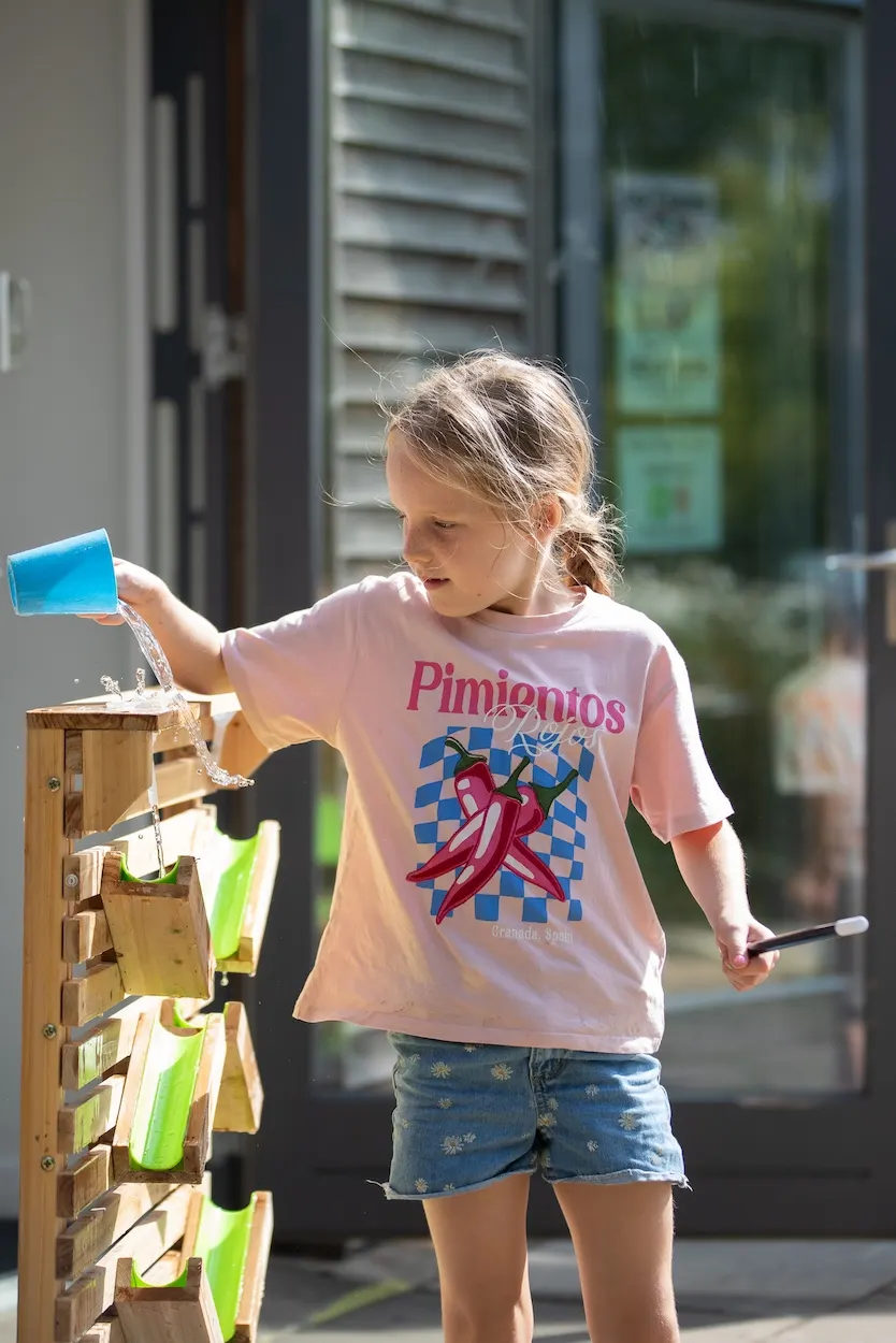 Young girl pouring water into a wooden water play feature in sunny outdoor courtyard.