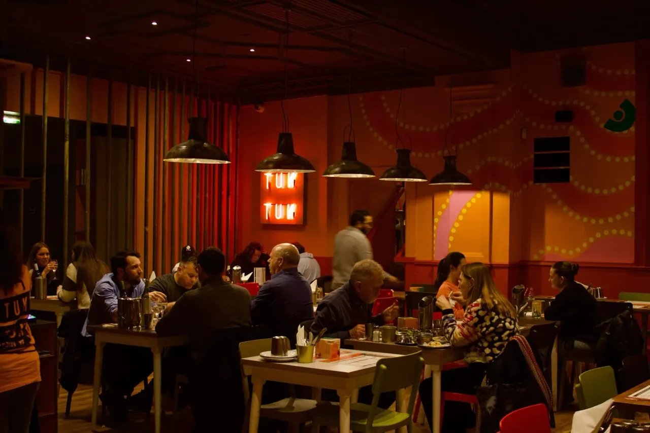 People dining and socialising in a vibrant red-lit restaurant with hanging pendant lights and neon signage.